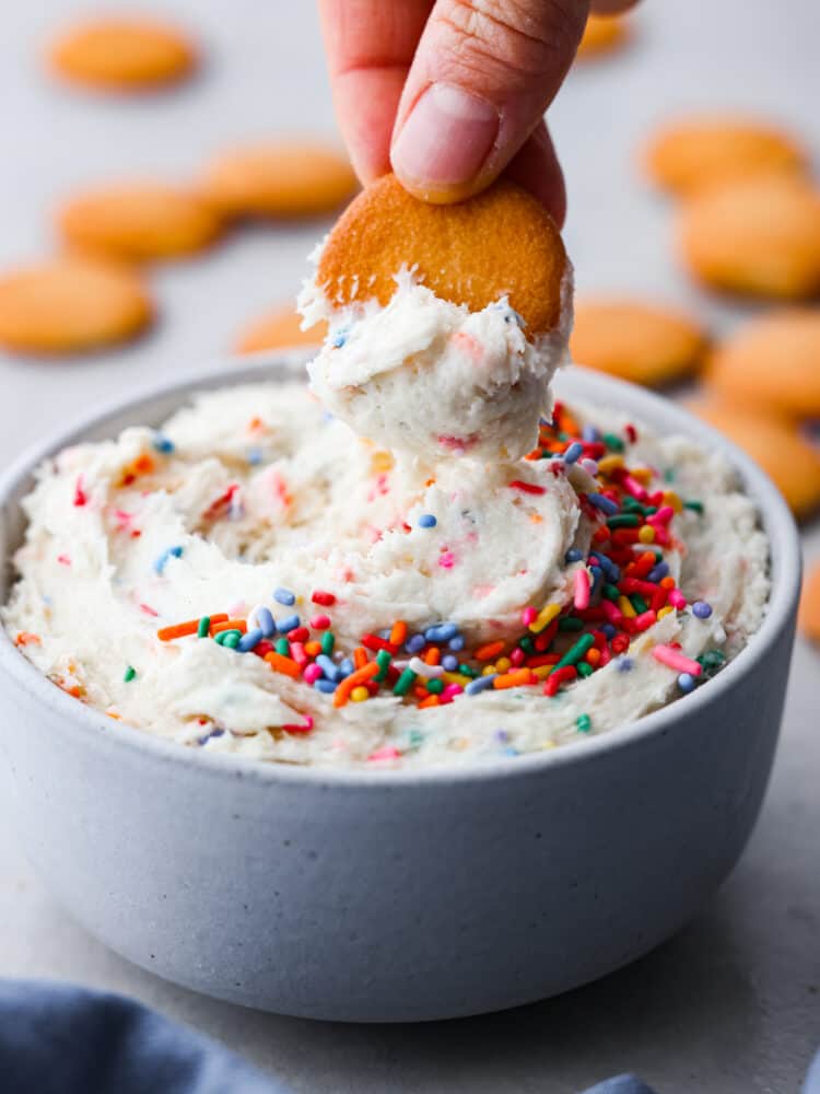 If you are looking for a quick and easy treat for a special occasion, this Dunkaroo Dip is your new go-to! It's a sweet dip perfect for cookies, apples, and graham crackers. It's colorful and absolutely addicting! A hand dipping a vanilla wafer cookie into dunkaroo dip.