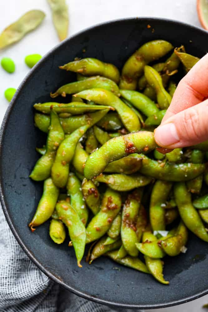 Garlic edamame is a tasty snack that's super simple to make. The combination of garlic, soy sauce, and flaky salt adds a delicious punch of flavor to the nutty edamame beans! It's a healthy, flavorful snack the whole family will enjoy. Picking up an edamame pod.