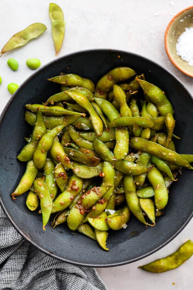 Garlic edamame is a tasty snack that's super simple to make. The combination of garlic, soy sauce, and flaky salt adds a delicious punch of flavor to the nutty edamame beans! It's a healthy, flavorful snack the whole family will enjoy. Top-down view of edamame served in a black bowl.