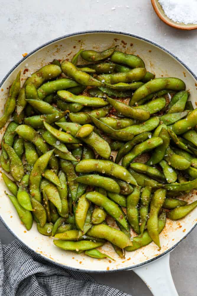 Garlic edamame is a tasty snack that's super simple to make. The combination of garlic, soy sauce, and flaky salt adds a delicious punch of flavor to the nutty edamame beans! It's a healthy, flavorful snack the whole family will enjoy. Garlic edamame in a blue and white skillet.