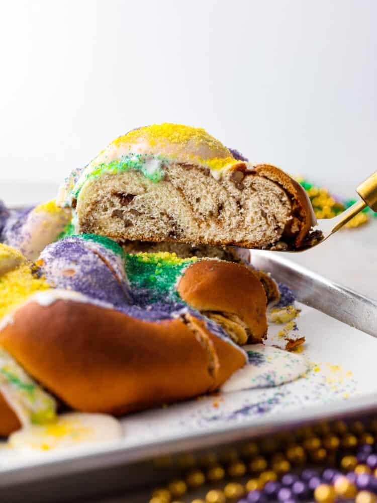 King cake is a colorful and festive dessert for Mardi Gras celebrations! With its gorgeous swirl of purple, green and gold icing, this sweet and soft cake is a treat for both the eyes and the taste buds. Whether you're a seasoned king cake aficionado or a newcomer to this sugary delight, there's no denying its irresistible charm! A slice of king cake being served with a spatula.