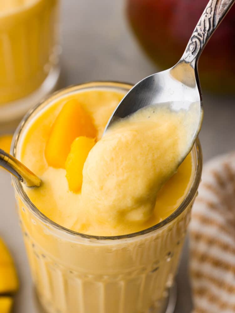 This Mango Lassi is an Indian yogurt smoothie that is so creamy and full of flavor. It's spiced with cardamom to give it a little kick, and it will have you coming back for seconds. It will be an instant hit every time you serve it! A spoon taking a scoop out of the smoothie.