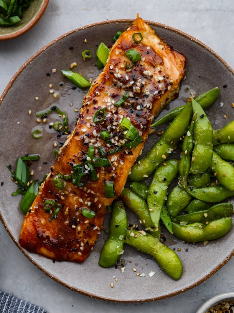 Better than at your favorite restaurant, this miso-glazed salmon is sweet, savory, and full of flavor. It cooks with a delicious caramelized miso glaze on the outside and stays flaky and moist in the middle. You are going to love everything about this recipe! A piece of salmon on a plate with edamame.