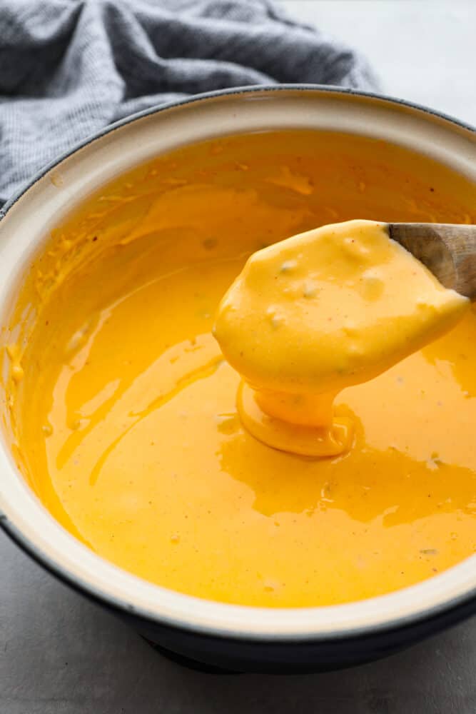 This Nacho Cheese recipe is the genuine Velveeta based topping that is full of nostalgia and amazing flavor! It's so simple to whip up and tastes way better than store-bought. Cheese sauce in a pot with a wooden spoon.