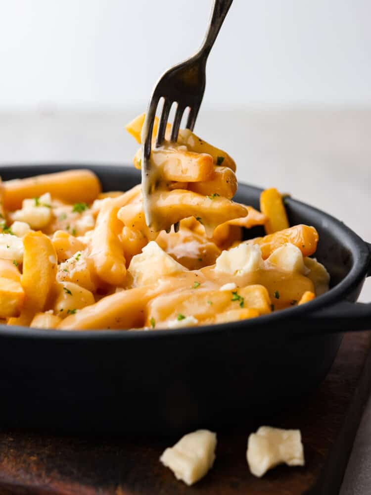 Poutine is a famous Canadian dish that you are going to love making at home! Delicious crispy fries topped with savory gravy and cheese curds are exactly what every party needs. You can serve them as an appetizer or side for any occasion! Poutine in a dish with a fork taking a piece of it.
