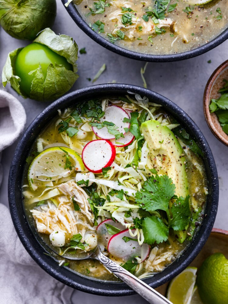 Pozole verde is a flavorful Mexican soup with fresh verde and shredded chicken. It is topped with ingredients like shredded cabbage, radishes, and lime for a bright, fresh, and flavor packed dish. It is the perfect soup for any occasion! Pozole verde in a black bowl with fresh ingredients around it.
