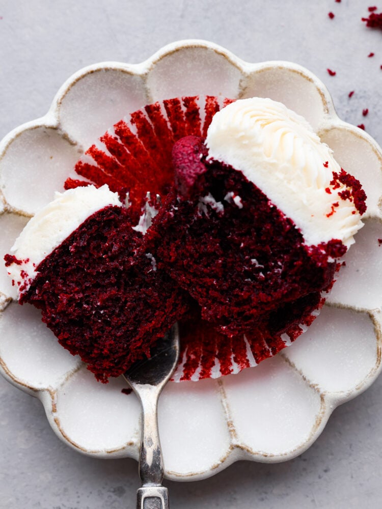 These red velvet cupcakes are perfectly moist cake with the most beautiful and deep red color. They are topped with classic cream cheese frosting, and once you eat one, you will agree that these are the best! A cupcake cut in half and served on a white plate.