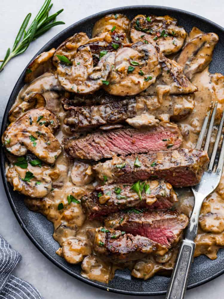 Juicy and succulent Steak Diane is the perfect dinner for any occasion. Diane refers to the delicious cream sauce made that includes mustard, Worcestershire sauce, and Cognac. The steak is flattened and seared in butter, and couldn't be more flavorful! Steak diane on a plate with strips of beef cut up and a fork to the side.