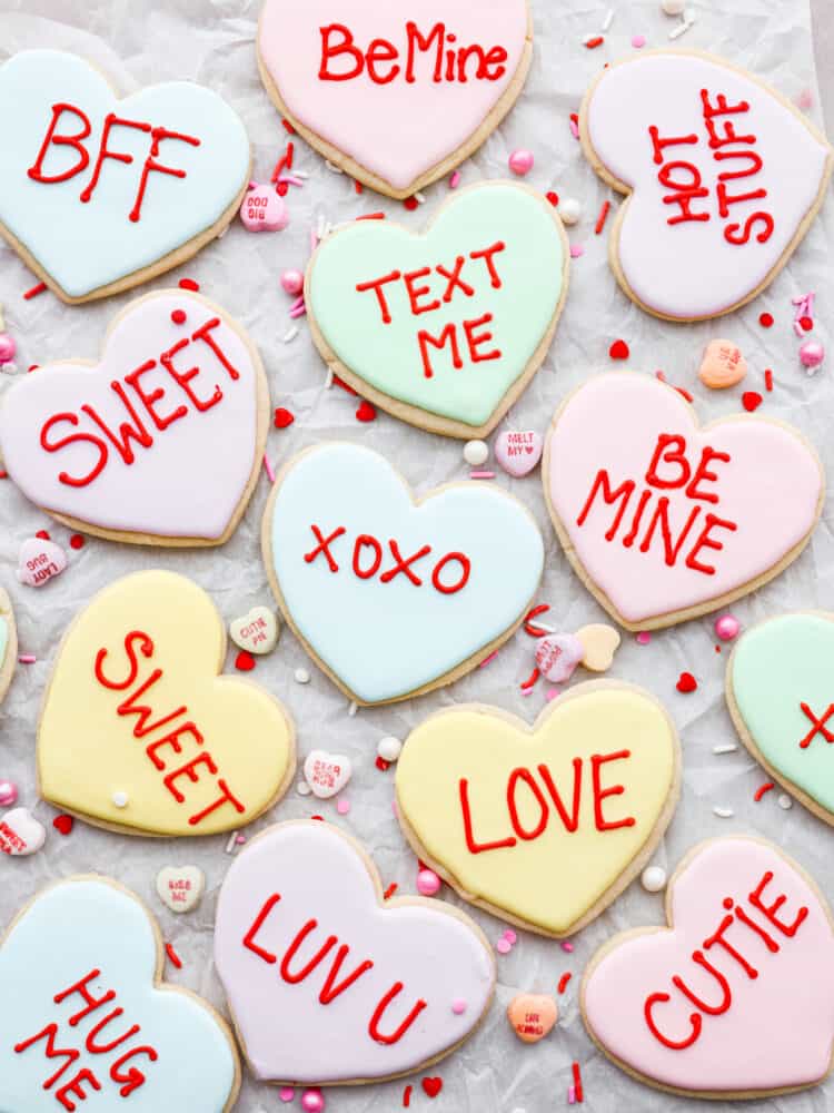 Show your sweetheart how much you care with these adorable Valentine's Day cookies! Conversation heart cookies are fun and nostalgic and are customizable with sweet and silly messages. They are delicious, easy to make, and totally unforgettable! An overhead view of all the Valentine's Day cookies on parchment paper with the conversation hearts next to them.