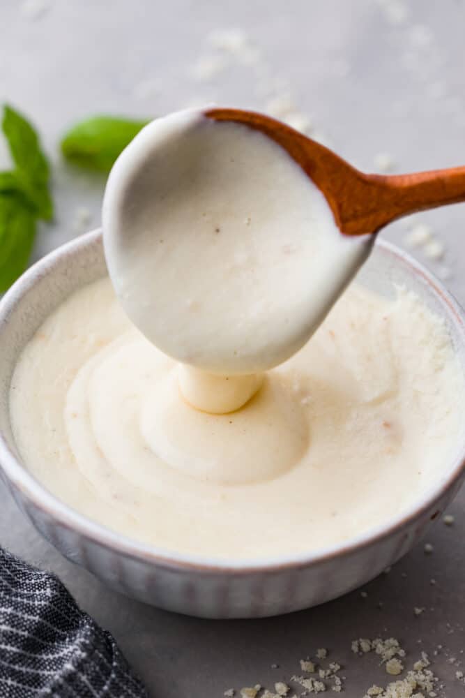 This creamy white pizza sauce is made with sharp parmesan cheese and rich half and half. It is so smooth and bursting with cheesy creamy flavors, and perfect on top of pizza, over bread, or for dipping your breadsticks in. White pizza sauce on a wooden spoon.