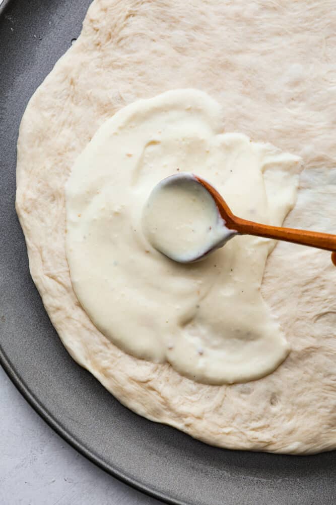 This creamy white pizza sauce is made with sharp parmesan cheese and rich half and half. It is so smooth and bursting with cheesy creamy flavors, and perfect on top of pizza, over bread, or for dipping your breadsticks in. A spoon spreading the sauce over pizza dough.