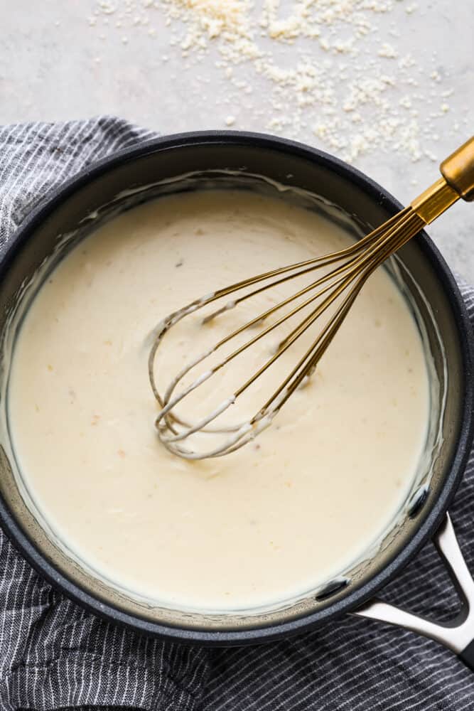 This creamy white pizza sauce is made with sharp parmesan cheese and rich half and half. It is so smooth and bursting with cheesy creamy flavors, and perfect on top of pizza, over bread, or for dipping your breadsticks in. A whisk in a bowl of the white pizza sauce.