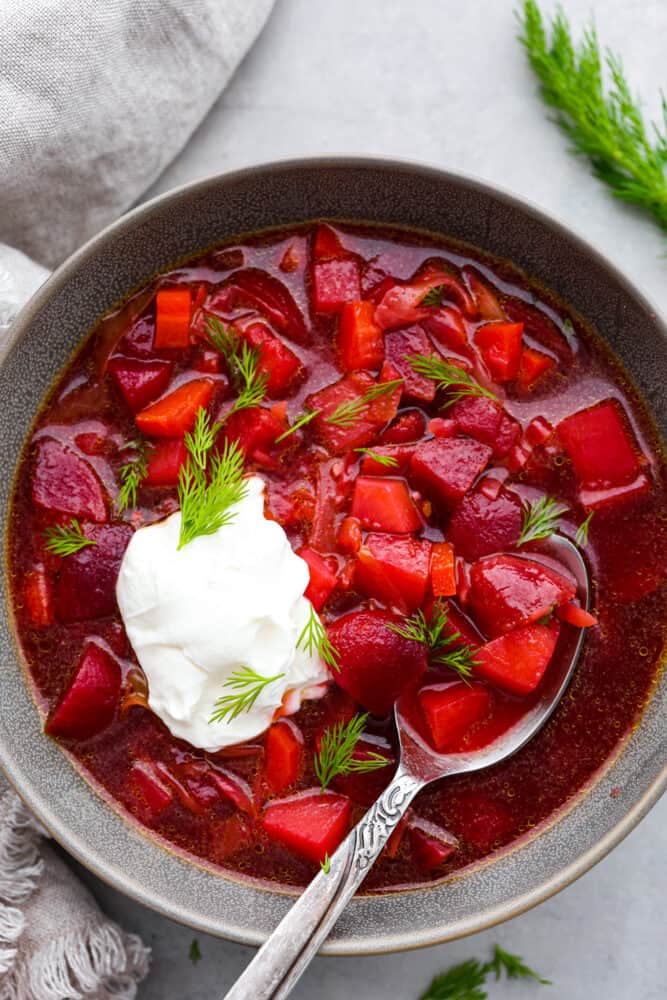 Borscht: the perfect soup for when you want to feel healthy but need something packed with flavor. It's like a hug in a bowl, if that hug was made of beets and potatoes. This tasty Ukrainian soup will become a new family favorite! Beet soup served in a gray bowl with a dollop of sour cream and some fresh dill on top.