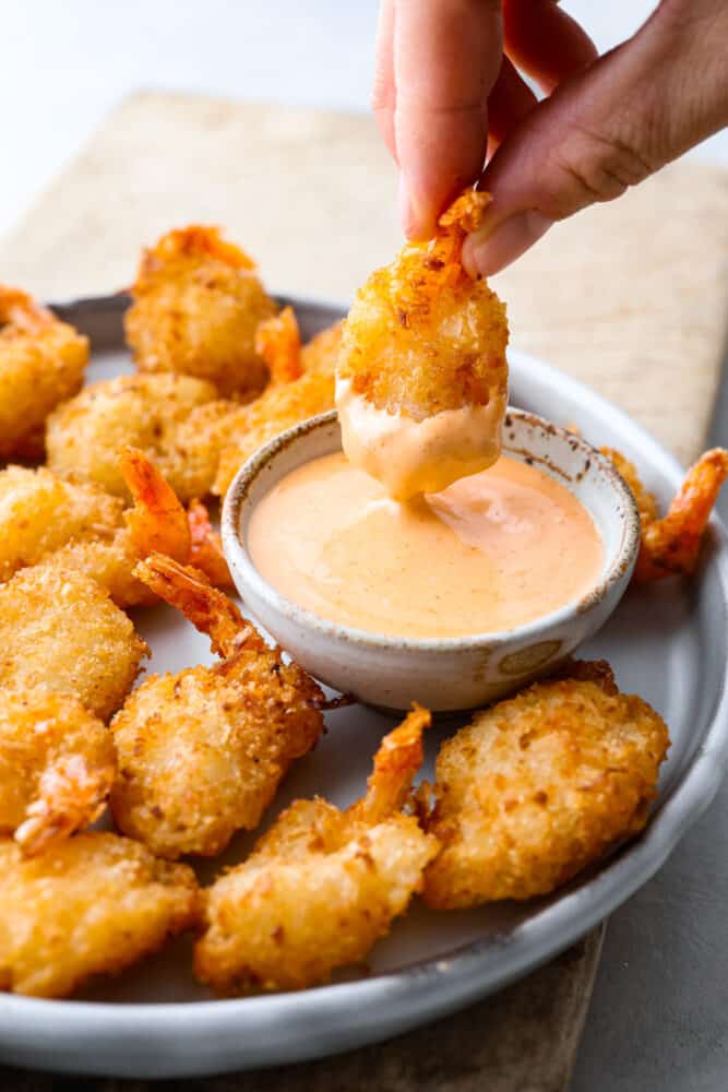 Seafood lovers! Here is the EASIEST way to make crispy, breaded shrimp in the air fryer! Making air fryer frozen shrimp takes less than 10 minutes and turns out perfectly every time. The hardest part is choosing what dipping sauce to serve it with! A piece of shrimp being dipped into a creamy sauce.