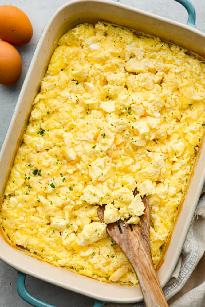 Get ready for the best scrambled eggs you've ever had (that also happen to be the easiest to make)! These Baked Scrambled eggs are light, fluffy, and full of flavor, and perfect for food prepping, holiday breakfasts, or just having enough food on the table to keep everyone full until lunch. Baked scrambled eggs in a casserole dish with a wooden spoon.