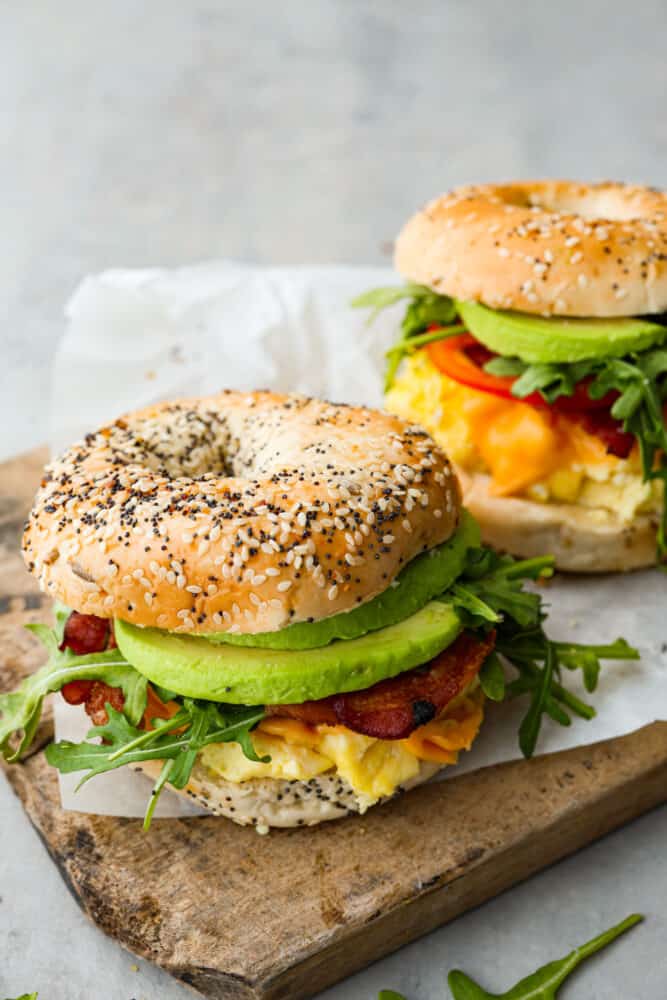 Start your morning off right with this hearty and filling Bagel Breakfast Sandwich. It has everything you need to fuel your body and to keep you full until lunch. These are also perfect for breakfast on the go and always hit the spot! Bagel breakfast sandwiches on a cutting board.