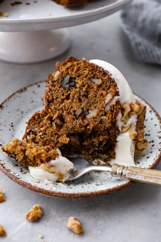Carrot bundt cake is a beautiful combination of moist and sweet carrot cake, topped with divine cream cheese frosting and chopped walnuts. It's full of warm spices and bright flavors that blend together for the perfect festive dessert. A piece of carrot bundt cake on a plate with a fork taking a scoop out.