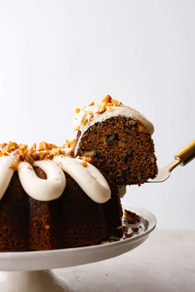Carrot bundt cake is a beautiful combination of moist and sweet carrot cake, topped with divine cream cheese frosting and chopped walnuts. It's full of warm spices and bright flavors that blend together for the perfect festive dessert. A slice of carrot bundt cake being lifted up by a spatula.