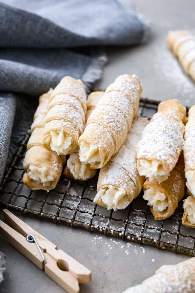 These clothespin cookies (also known as cream horns) are like little bites of heaven, filled with creamy goodness that'll make you forget all your problems - at least until you run out of them. They only take 30 minutes to prepare and are just the soft, creamy pastry you need in your life! Multiple clothespin cookies stacked on top of each other on a cooling rack.
