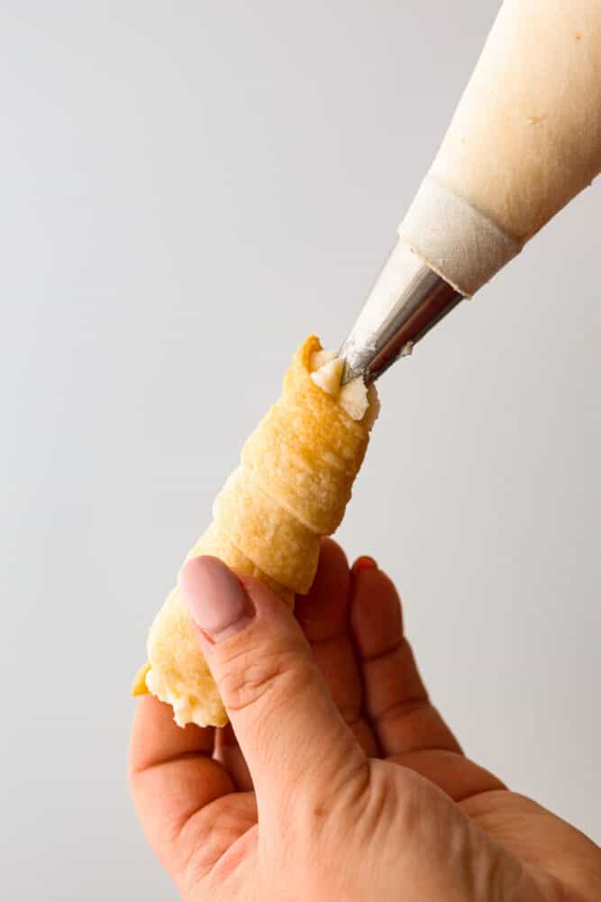 These clothespin cookies (also known as cream horns) are like little bites of heaven, filled with creamy goodness that'll make you forget all your problems - at least until you run out of them. They only take 30 minutes to prepare and are just the soft, creamy pastry you need in your life! A cream horn cookie shell being piped with filling.