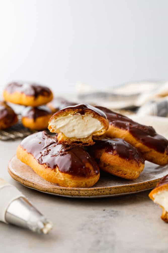 Skip the bakery and make your own delicious eclairs at home! These cream-filled delights are a labor of love and are sure to become one of your new favorite dessert recipes. 4 eclairs on a plate. One is cut in half.