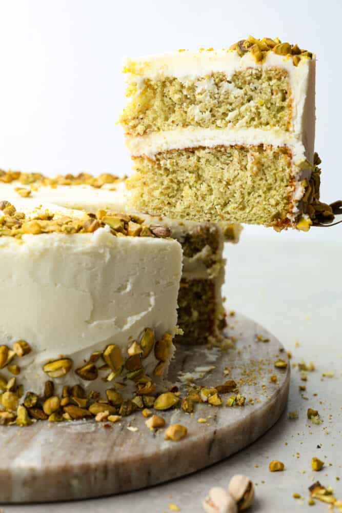 This light and airy Pistachio Cake is made from scratch, full of real pistachios, and topped with a delicious buttercream frosting. It's a classy cake that will impress your guests on any occasion! Pistachio cake slice being lifted up with a spatula.