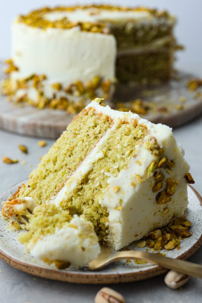 This light and airy Pistachio Cake is made from scratch, full of real pistachios, and topped with a delicious buttercream frosting. It's a classy cake that will impress your guests on any occasion! A slice of pistachio cake on a plate.