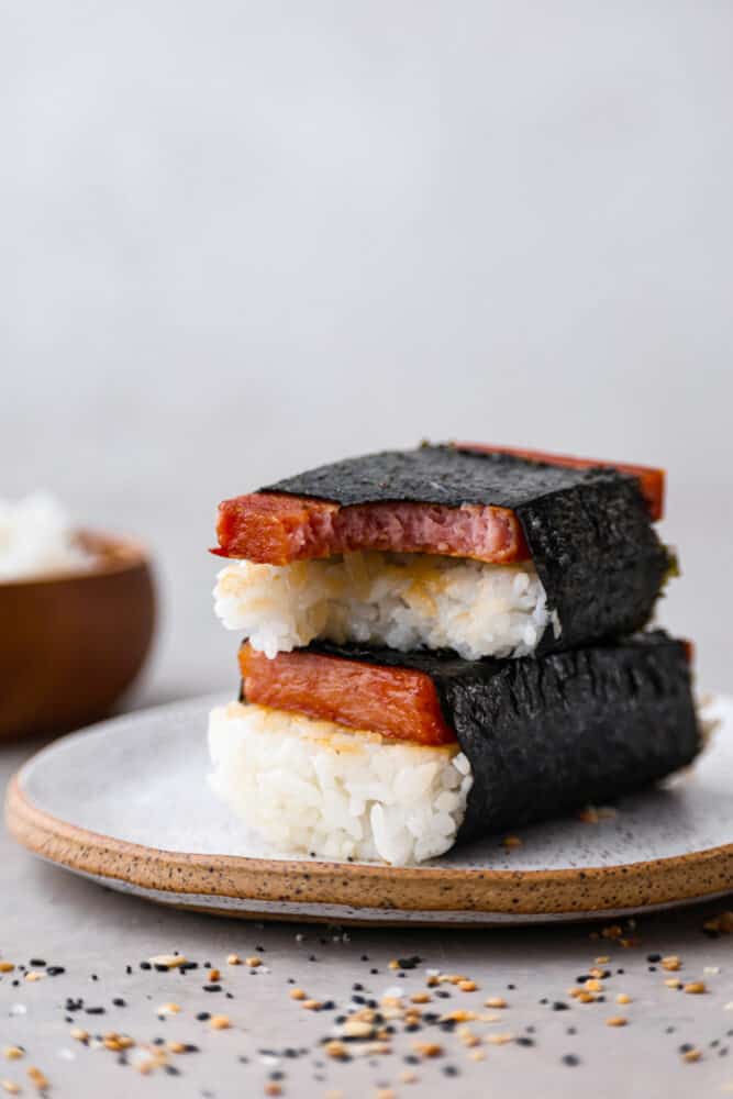 Spam musubi is a classic Hawaiian snack or on-the-go lunch, made with a slice of seared spam on top of rice, all wrapped in crispy seaweed! It has a delicious soy sauce glaze that makes each bite extra flavorful. 2 musubi on a stoneware plate. One has a bite taken out of it.