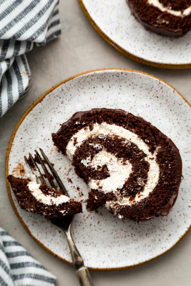 A classic and beautiful dessert, this Swiss Roll Cake is a rich chocolate sponge filled with pillowy whipped cream and covered in a chocolate ganache. This dessert is not only stunning, it's also incredibly tasty and every chocolate lover's dream! A piece of cake on the plate with a bite taken out of it.
