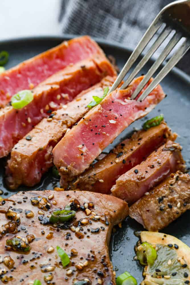 There's no reason to save Tuna Steaks for eating out, make them yourself for a gourmet restaurant experience in the comfort of your home. Not only are they full of healthy nutrients, they are delicious and a real treat any day of the week. A close up on the dish with a fork taking a slice.