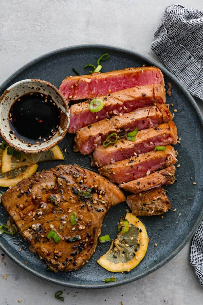 There's no reason to save Tuna Steaks for eating out, make them yourself for a gourmet restaurant experience in the comfort of your home. Not only are they full of healthy nutrients, they are delicious and a real treat any day of the week. Seared ahi tuna steak on a plate cut up with a dipping sauce.