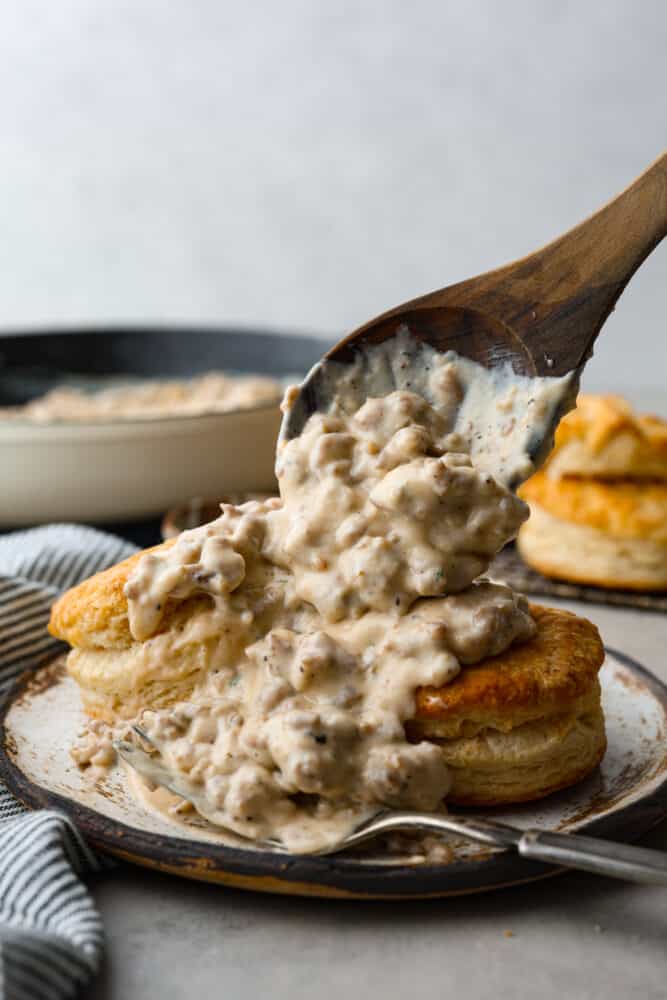 Biscuits and Gravy is the ultimate comfort food dish! Tender and flaky homemade biscuits are topped with creamy sausage gravy. This is such a simple recipe that is full of rich and nostalgic flavors, you've got to try it! Biscuits and gravy on a plate with a wooden spoon adding more gravy to the biscuits.