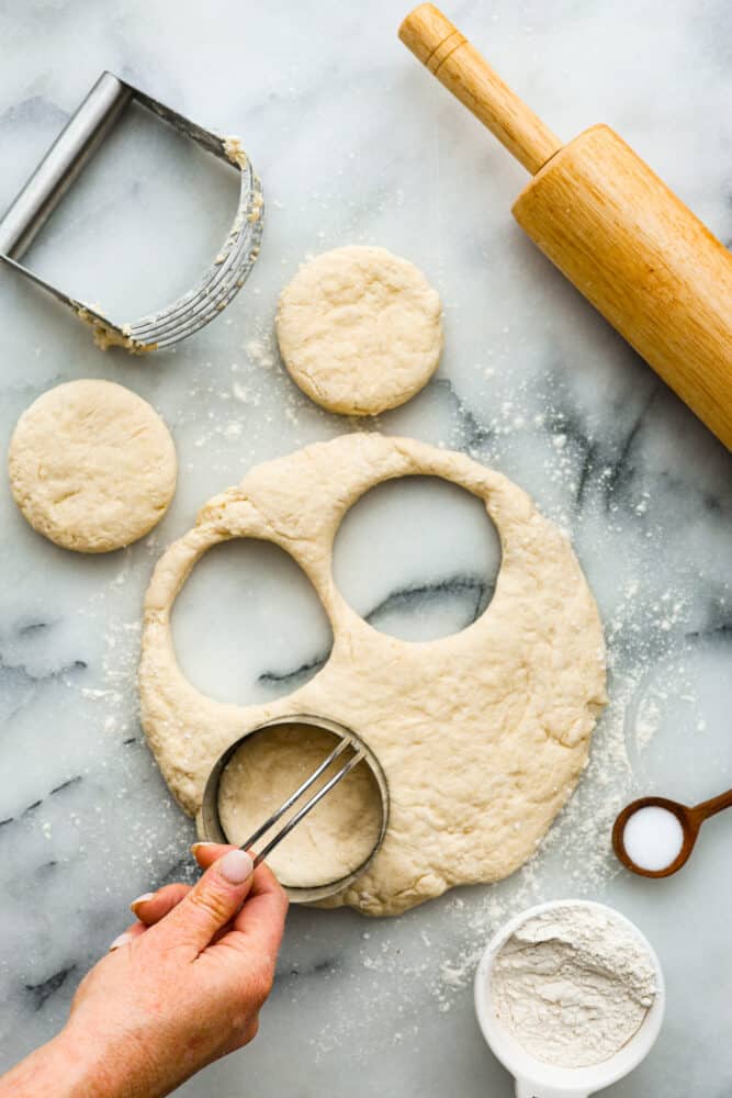 Biscuits and Gravy is the ultimate comfort food dish! Tender and flaky homemade biscuits are topped with creamy sausage gravy. This is such a simple recipe that is full of rich and nostalgic flavors, you've got to try it! A hand holding a biscuit cutter cutting the biscuit dough.