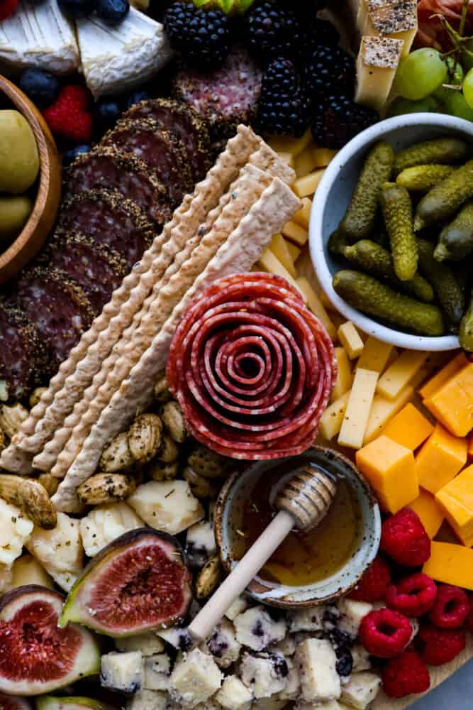 Charcuterie boards are the ultimate party trick that never fail to impress! These beautiful displays of meats, cheeses, fruits, and nuts are not only eye-catching but also incredibly delicious. Don't be intimidated by how Pinterest-worthy they look, because assembling one is a lot easier than you think. Here are all the ins and outs on how to make a charcuterie board at home! A salame rose surrounded by things like pickles, cubed cheese, honey, and crackers.