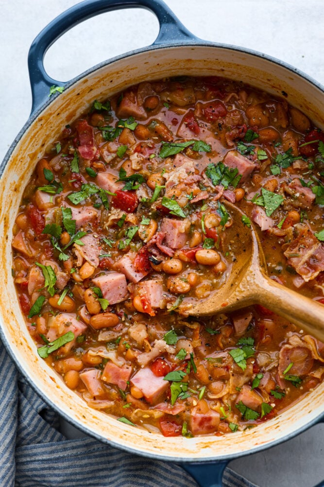 Mexican Charro Beans, also known as Frijoles Charros, are loaded to the brim with flavor! Pinto beans are cooked in a chicken broth seasoned with warm and inviting spices. Throw in some bacon, tomato, onions, ham, and jalapeños for the perfect blend of bold flavors. Charro beans in a pot with a wooden spoon.