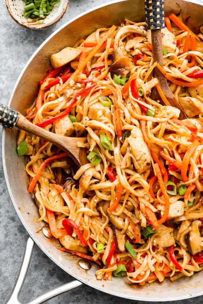 Homemade Chicken Lo Mein is a classic Chinese noodle dish that tastes so much better than takeout! Noodles drenched in a delicious savory sauce paired with perfectly cooked chicken and tender peppers and carrots makes for a yummy and satisfying meal. A close up on chicken lo mein in the skillet with a wooden spoon in it.