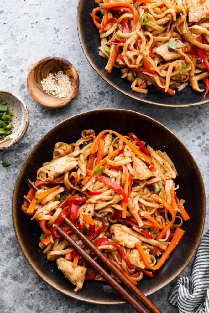 Homemade Chicken Lo Mein is a classic Chinese noodle dish that tastes so much better than takeout! Noodles drenched in a delicious savory sauce paired with perfectly cooked chicken and tender peppers and carrots makes for a yummy and satisfying meal. Chicken lo mein in a black bowl with other bowls around it.