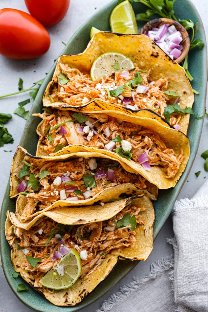 If you're a fan of Mexican cuisine, you have to try chicken tinga! This delicious dish is a classic from the Puebla region and is made with shredded chicken that's been simmered in a flavorful tomato-based sauce with smoky chipotle peppers. The result is a savory, slightly spicy, and oh-so-tasty dish that's perfect for tacos, tostadas, or just served over rice. Trust me, once you taste it, you'll be hooked! Top-down view of 4 chicken tacos on a green plate. They are topped with chopped onions, cheese crumbles, and cilantro.