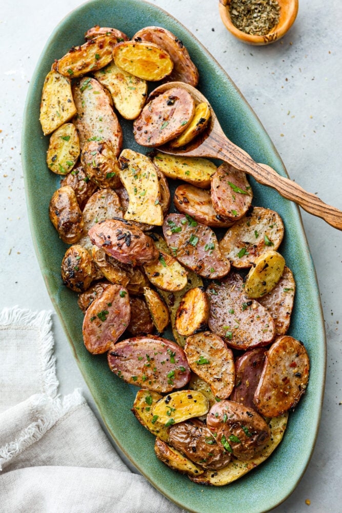 Roasted Fingerling Potatoes are thin skinned and crispy on the outside, yet so creamy and smooth on the inside. They are smaller potatoes, so they cook quickly and are loaded with fresh seasonings that make these potatoes irresistible. Fingerling potatoes on a plate with a wooden spoon.