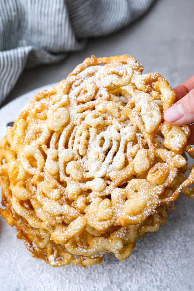 Skip the fair and make your own funnel cake at home! The batter is so quick and easy, you only need 20 minutes to make this fun treat at home. It will feel like a night out and taste better than any funnel cake you've ever had. A hand grabbing a funnel cake.