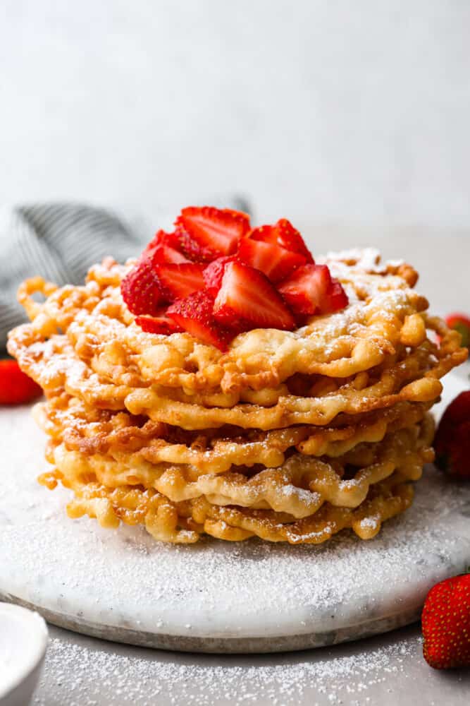 Skip the fair and make your own funnel cake at home! The batter is so quick and easy, you only need 20 minutes to make this fun treat at home. It will feel like a night out and taste better than any funnel cake you've ever had. Funnel cakes stacked on top of each other topped with strawberries and powdered sugar.