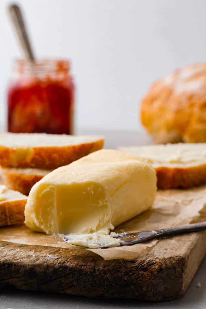 Storebought butter can be full of unnecessary and lower quality ingredients- make your own butter at home instead! You will be blown away by how flavorful and delicious this homemade butter is. Not to mention it's a really fun activity for the whole family too! Homemade butter on a cutting board with bread in the background.