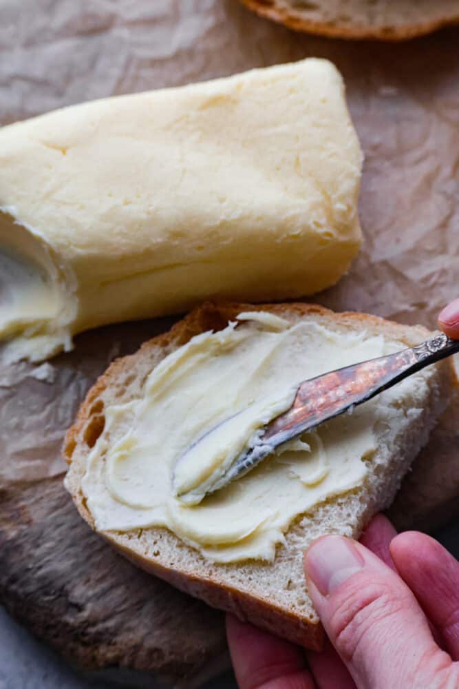 Storebought butter can be full of unnecessary and lower quality ingredients- make your own butter at home instead! You will be blown away by how flavorful and delicious this homemade butter is. Not to mention it's a really fun activity for the whole family too! A hand spreading butter onto a piece of bread with a butter knife.
