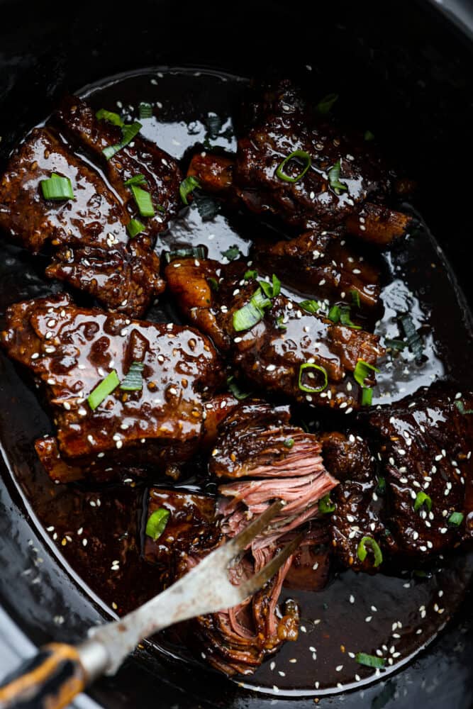 These bone-in Korean short ribs are sticky savory perfection! This recipe is as simple as searing your ribs and tossing them in a marinade and into the crock pot. The result is tender and flavorful meat that will have you coming back for seconds and thirds! A close up on the short ribs in the slow cooker being pulled apart.