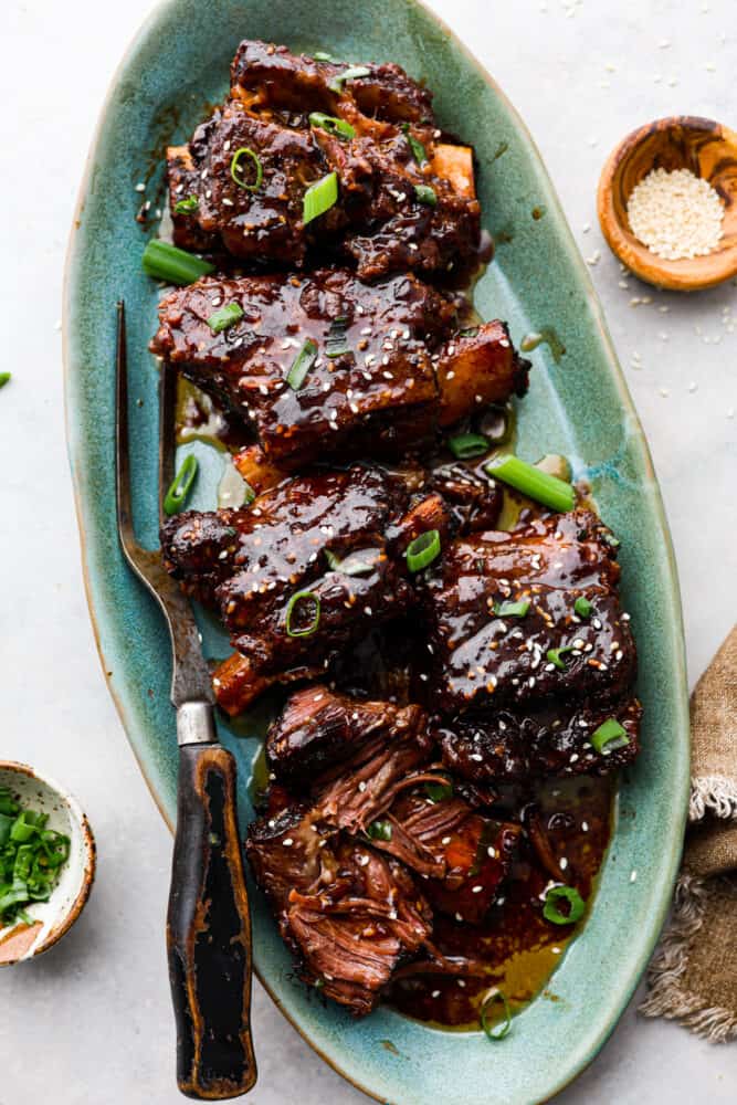 These bone-in Korean short ribs are sticky savory perfection! This recipe is as simple as searing your ribs and tossing them in a marinade and into the crock pot. The result is tender and flavorful meat that will have you coming back for seconds and thirds! Korean short ribs on a platter with tongs.