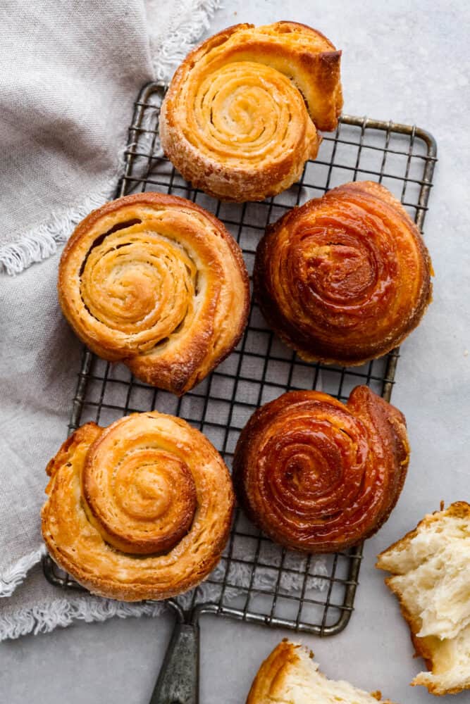 Imagine a sticky bun and a croissant put together and you have a delicious Kouign Amman! It's a laminated sweet dough that you will absolutely fall in love with. Don't be intimidated by this recipe, I break it down step by step into simple to follow instructions that will turn out great every single time! Kouign amman on a wire rack with a grey hand towel.