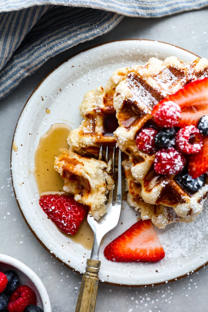 You've never had a waffle like a Liege Waffle! These waffles are similar to a Belgian waffle but made sweeter by pearl sugar. They are perfectly crispy on the outside and warm and soft on the inside. Topped with fresh berries and whipped cream, you won't find a better brunch companion! A liege waffle on a plate with a bite being scooped out with a fork.