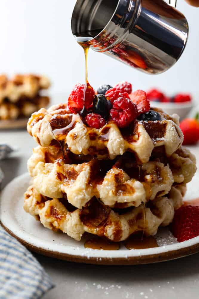 You've never had a waffle like a Liege Waffle! These waffles are similar to a Belgian waffle but made sweeter by pearl sugar. They are perfectly crispy on the outside and warm and soft on the inside. Topped with fresh berries and whipped cream, you won't find a better brunch companion! Liege waffles stacked on top of each other, topped with berries, syrup being poured on top.