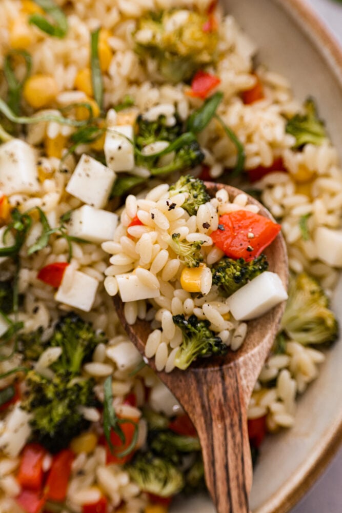 This Orzo Pasta Salad is filled with fresh veggies and zesty lemon flavor that will excite your tastebuds. It's perfect for your next potluck, party, or a refreshing and unique meal this summer. You're going to want to save this recipe for later! Orzo pasta salad on a wooden spoon with a bowl in the background.