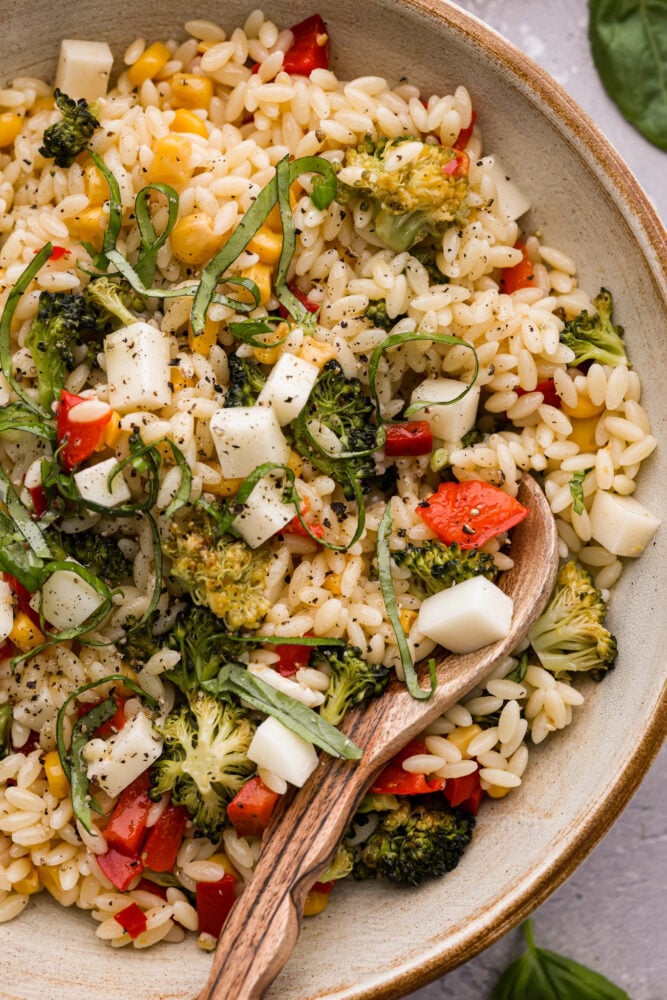 This Orzo Pasta Salad is filled with fresh veggies and zesty lemon flavor that will excite your tastebuds. It's perfect for your next potluck, party, or a refreshing and unique meal this summer. You're going to want to save this recipe for later! A close up on orzo pasta salad with a wooden spoon.